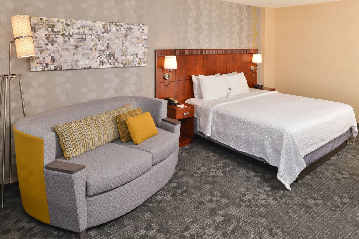 Habitación de hotel con un sofá gris, una cama con ropa blanca, cabecera de madera y una gran obra de arte abstracto en la pared.