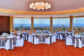 Un salón de banquetes con mesas redondas, sillas blancas y vistas a montañas a través de amplios ventanales.