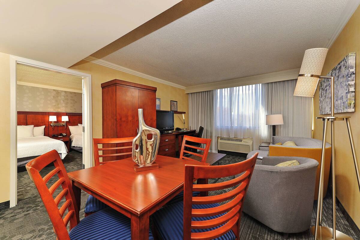 Habitación de hotel con una mesa de comedor de madera, dos sillas de madera, un sillón gris, un pequeño sofá, un escritorio con lámpara, un televisor y una cama con ropa de cama blanca.