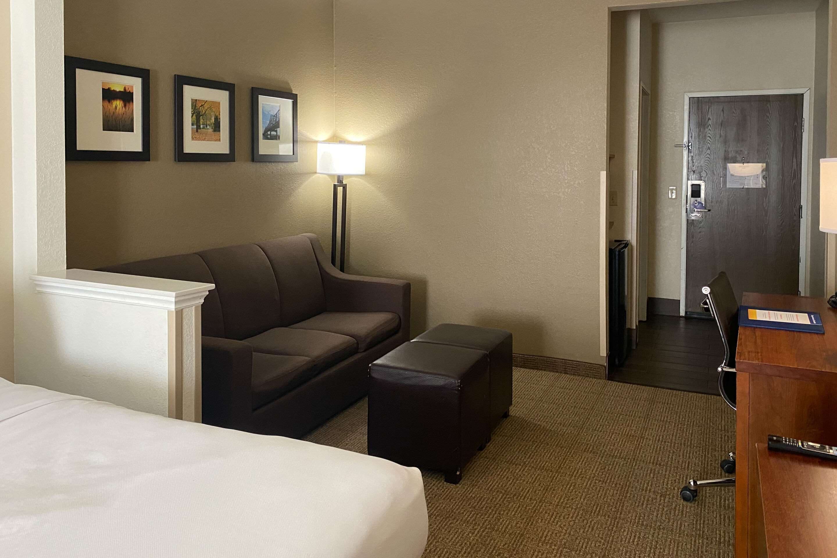 A hotel room with a bed, a sofa, a small table, a desk, and a lamp. There are framed pictures on the wall and a door leading to another room.