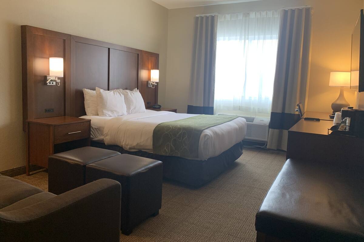 Ein Hotelzimmer mit Bett, Nachttischen, einem Schreibtisch, einem Sessel und einem großen Fenster mit Vorhängen.