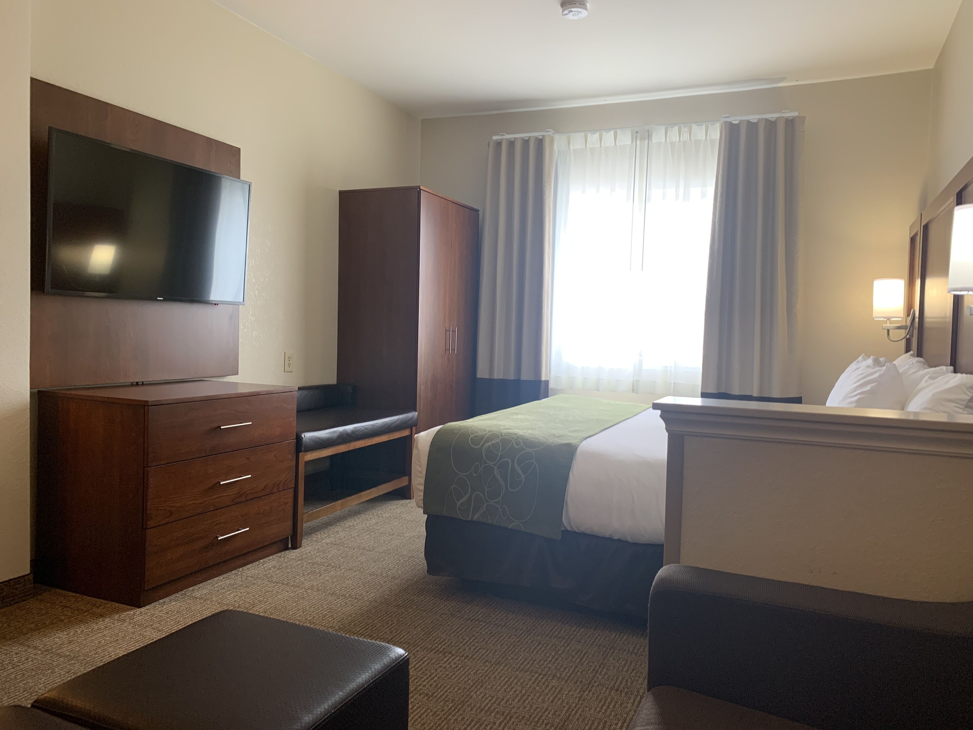 A hotel room with a bed, a TV mounted on the wall, a wardrobe, a desk, and a window with curtains.