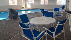 Zona de piscina con sillas azules y mesas blancas, piscina techada y ventanas grandes.