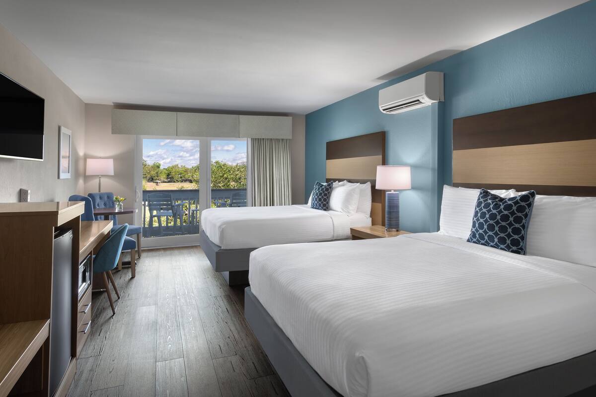 A hotel room with two beds, a desk, a chair, a TV, and a balcony with a view.