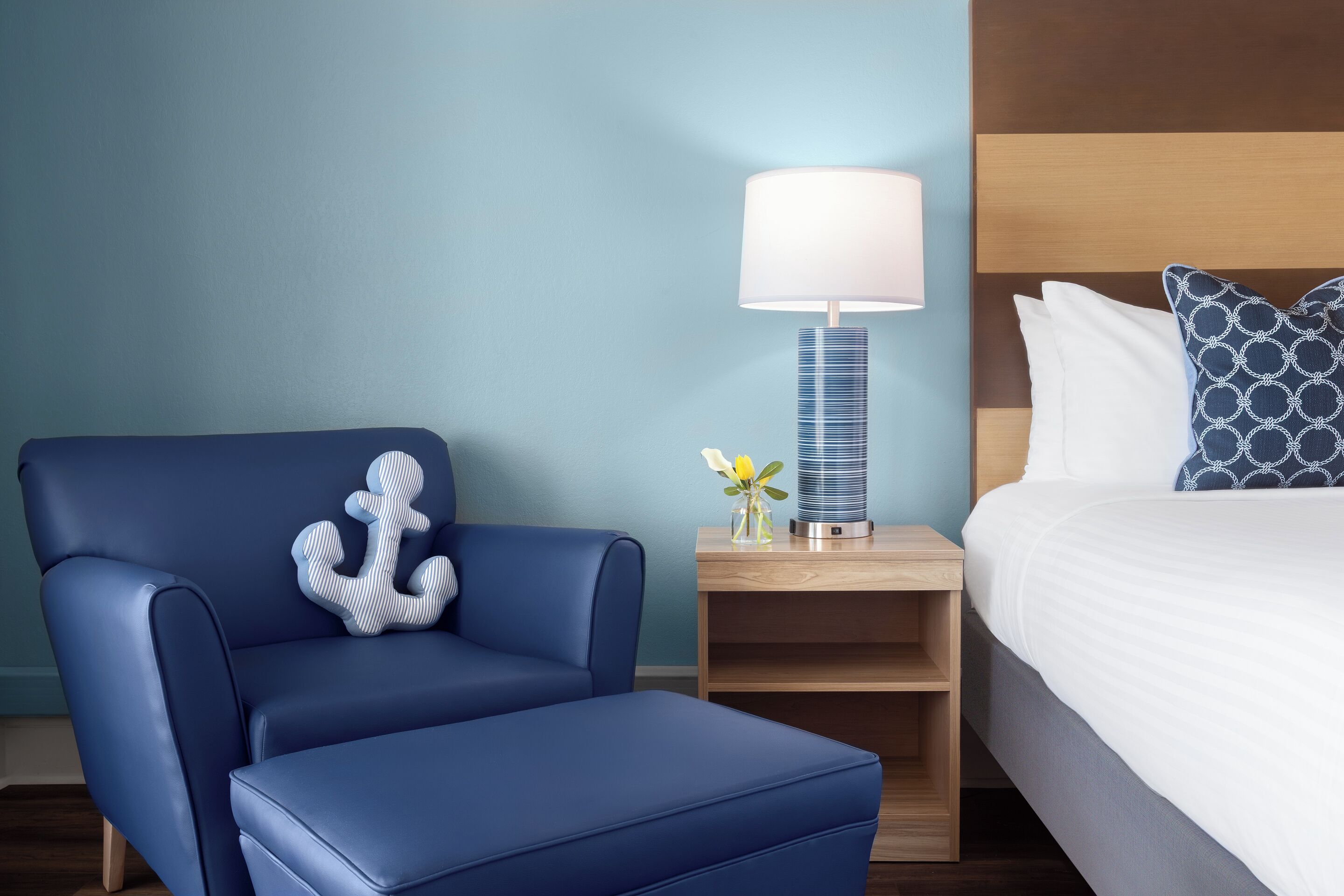 Een hotelkamer met een blauwe fauteuil, een nachtkastje met een lamp en een bed met gekleurde kussens.