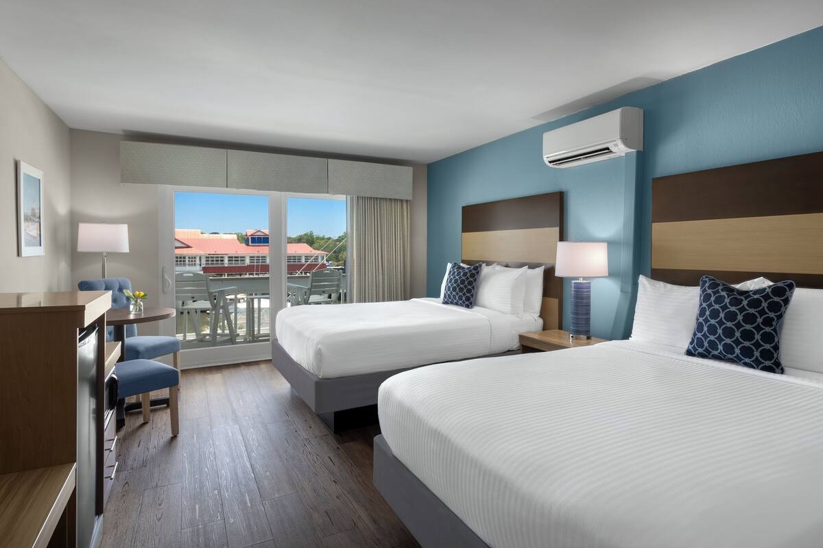 A hotel room with two beds, a desk, a chair, and a balcony with a view of buildings.
