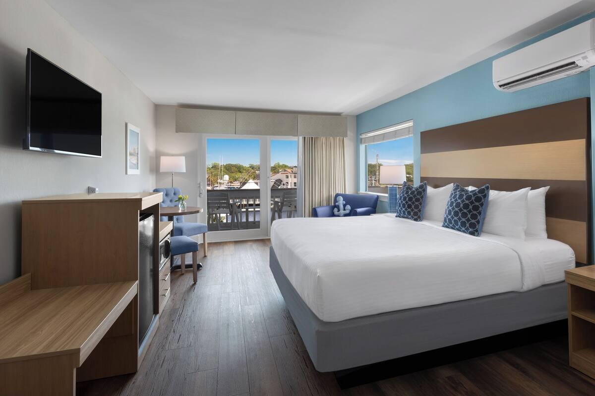 A hotel room with a large bed, a desk, a chair, a TV, and a balcony with a view of boats.