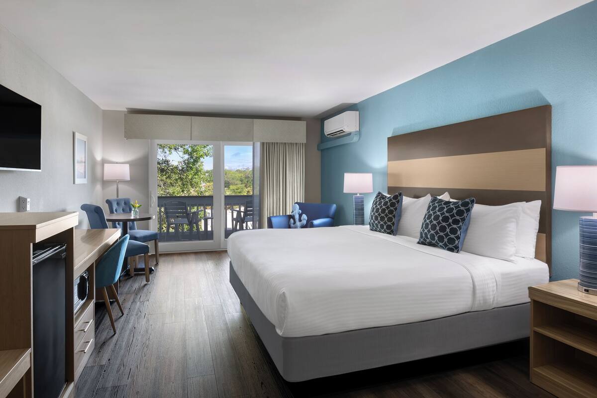A hotel room with a large bed, a desk, a chair, and a balcony with a view of trees.