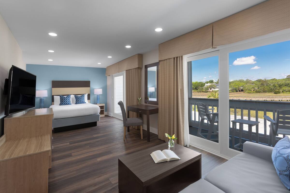 A modern hotel room with a bed, a desk, a TV, and a balcony with outdoor furniture.