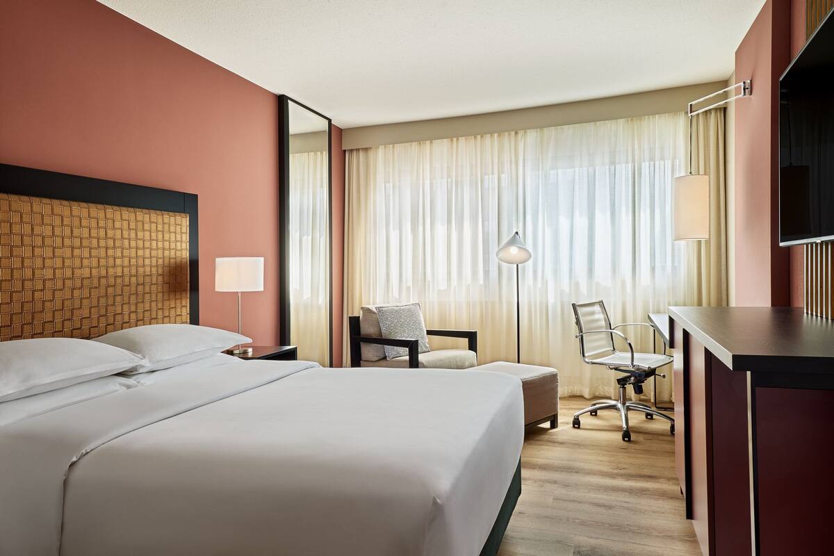 A hotel room with a large bed, a desk, a chair, a lamp, and a TV.