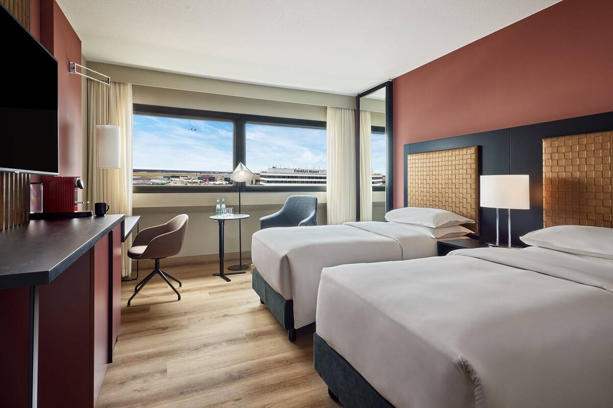 A hotel room with two beds, a desk, a chair, and a view of the airport.