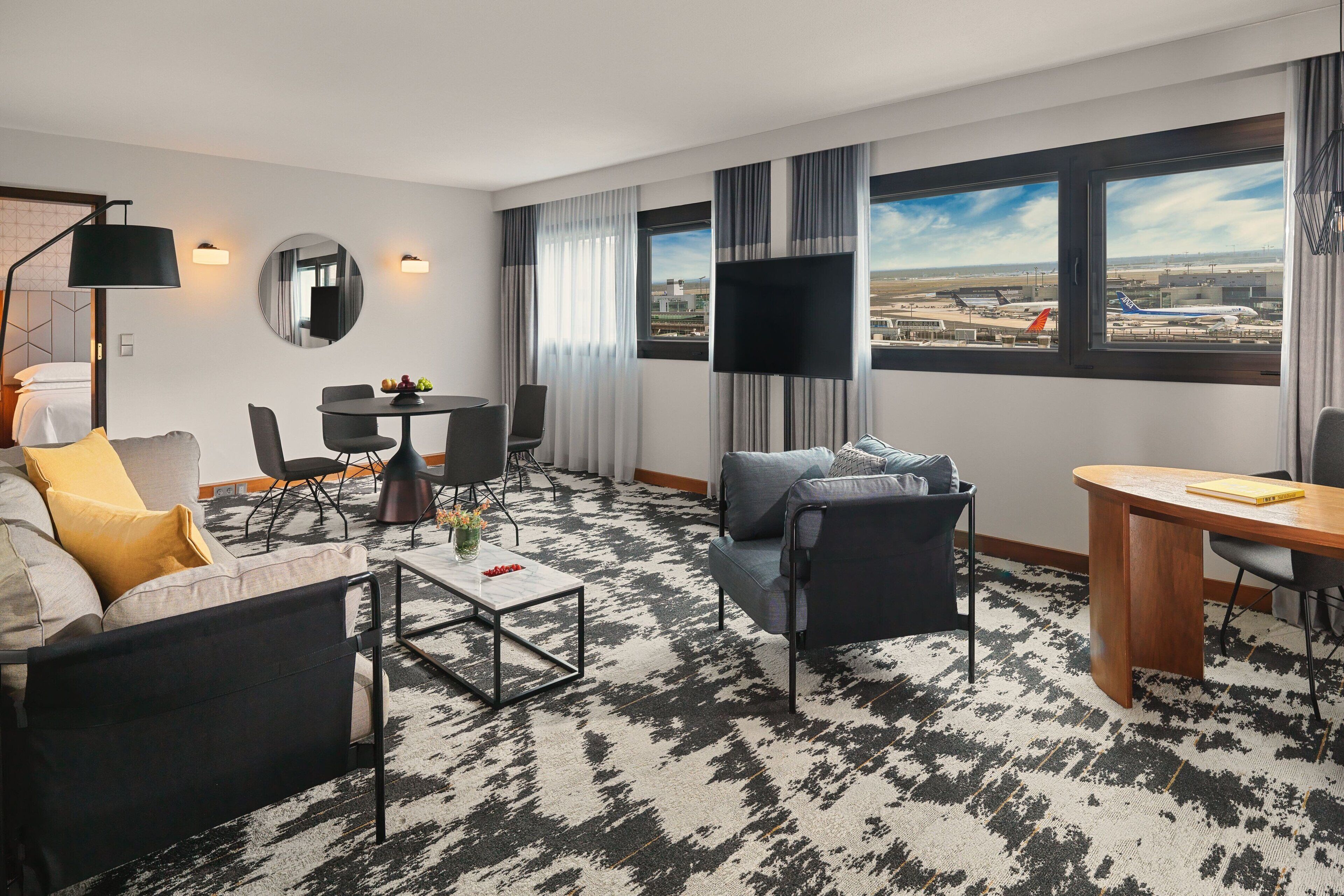 Une chambre d’hôtel moderne équipée d’un canapé, d’un fauteuil, d’une table à manger et d’une grande fenêtre donnant sur l’aéroport.