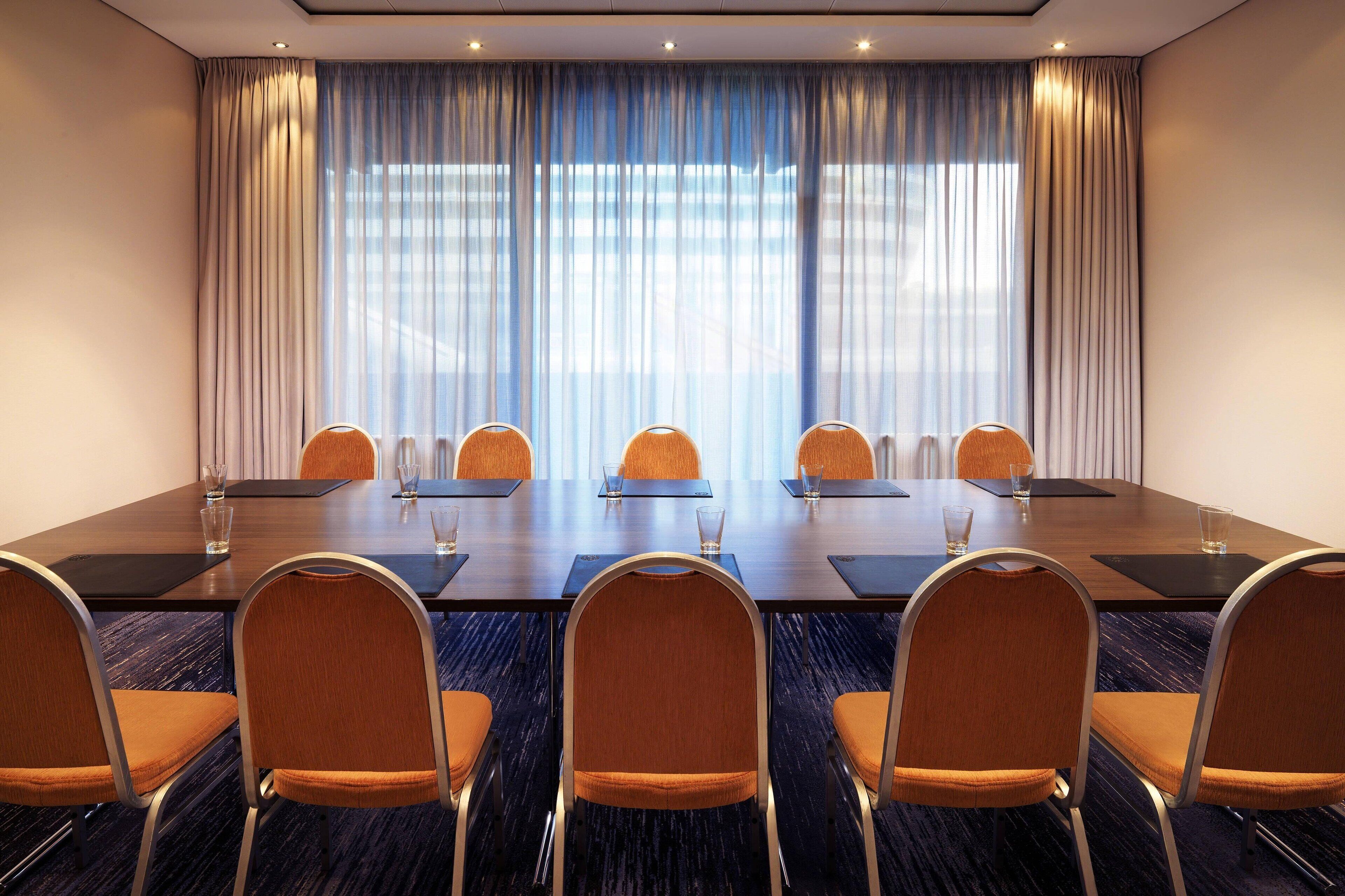 A conference room with a long table, six chairs, and a large window with curtains.