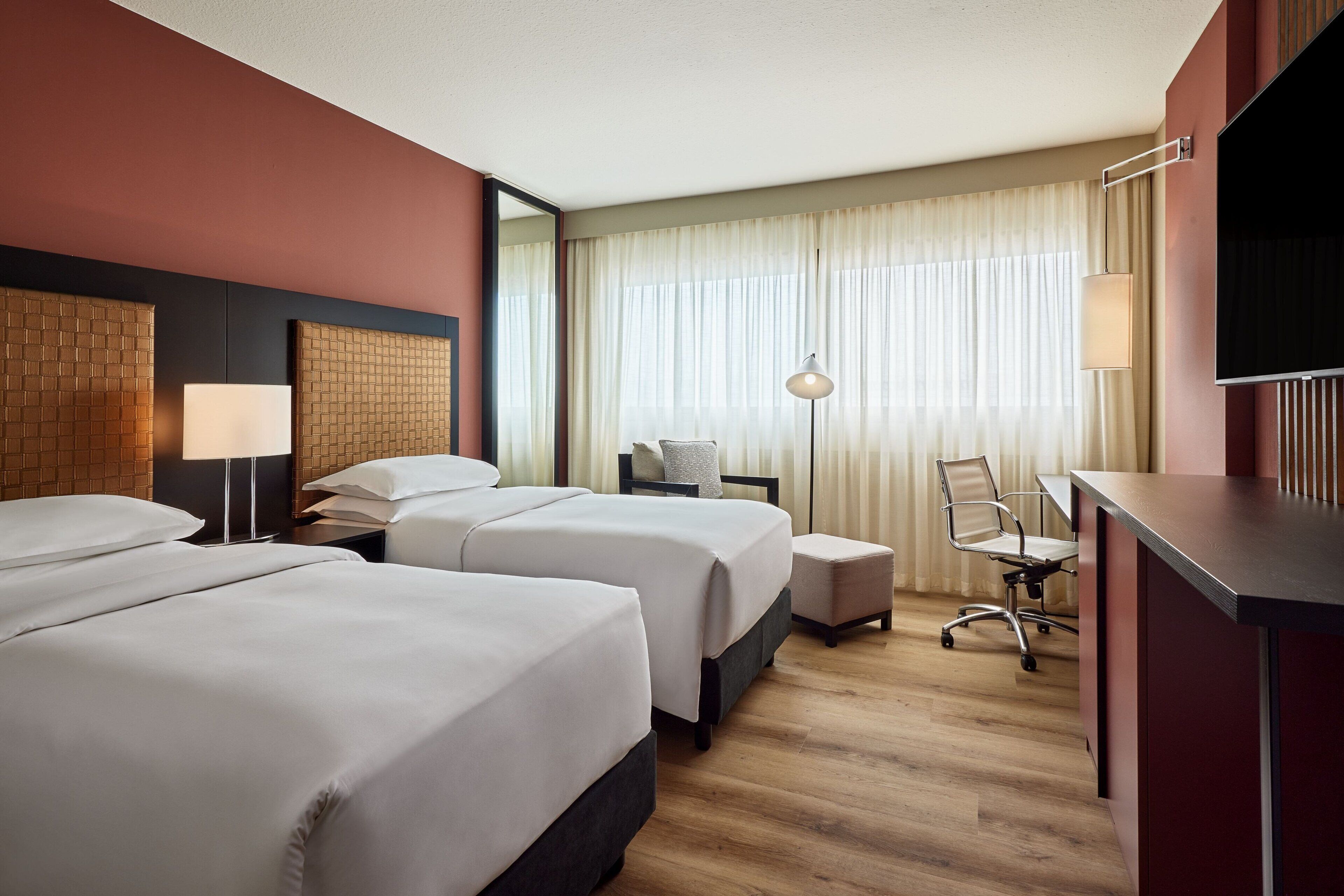 Ein Hotelzimmer mit zwei Betten, einem Schreibtisch, einem Stuhl, einem Fernseher und einem großen Fenster mit Vorhängen.