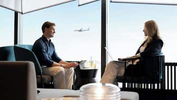 Ein modernes Hotelzimmer mit großen Fenstern, einem Laptop, einem Glas Wasser und einer Zeitschrift auf dem Tisch.