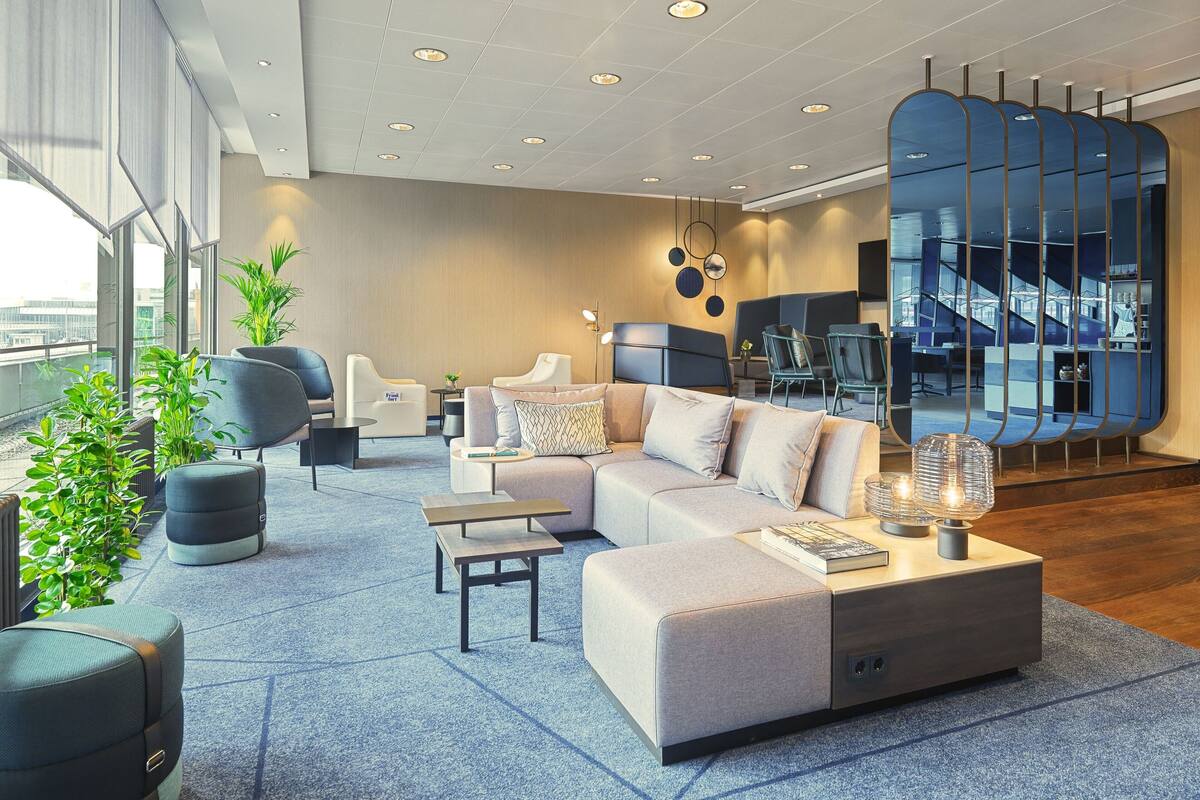 A modern hotel lobby with a large sectional sofa, a round coffee table, and a seating area with chairs. There is a staircase with a glass railing visible in the background.