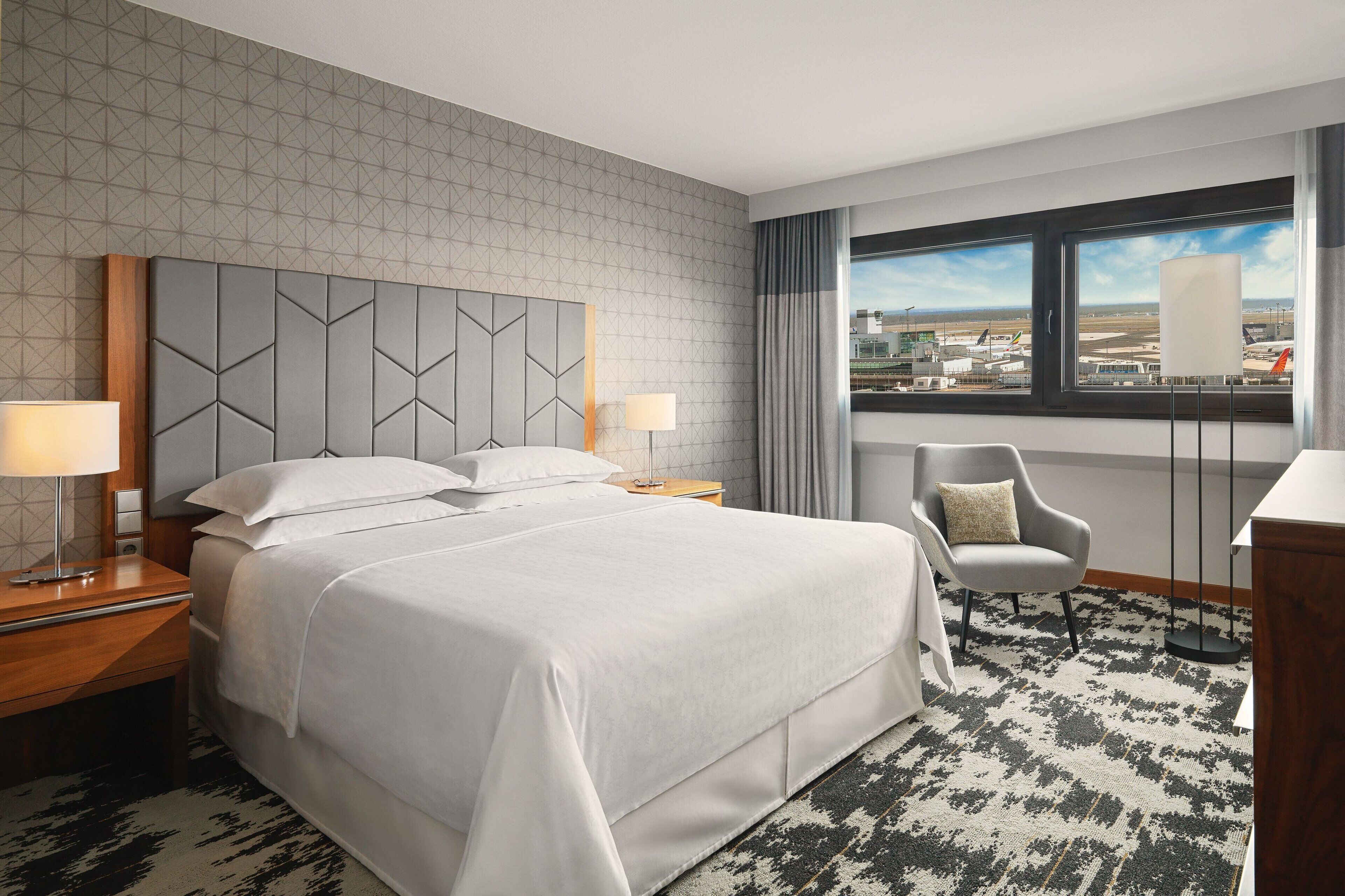 A hotel room with a large bed, two bedside tables with lamps, a chair, a window with a view of the airport, and a patterned carpet.