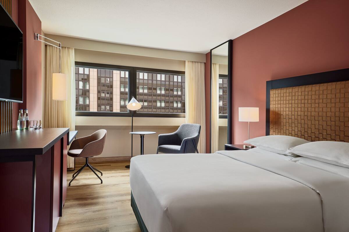 A hotel room with a large bed, a desk with a chair, a small table, and a window with curtains.