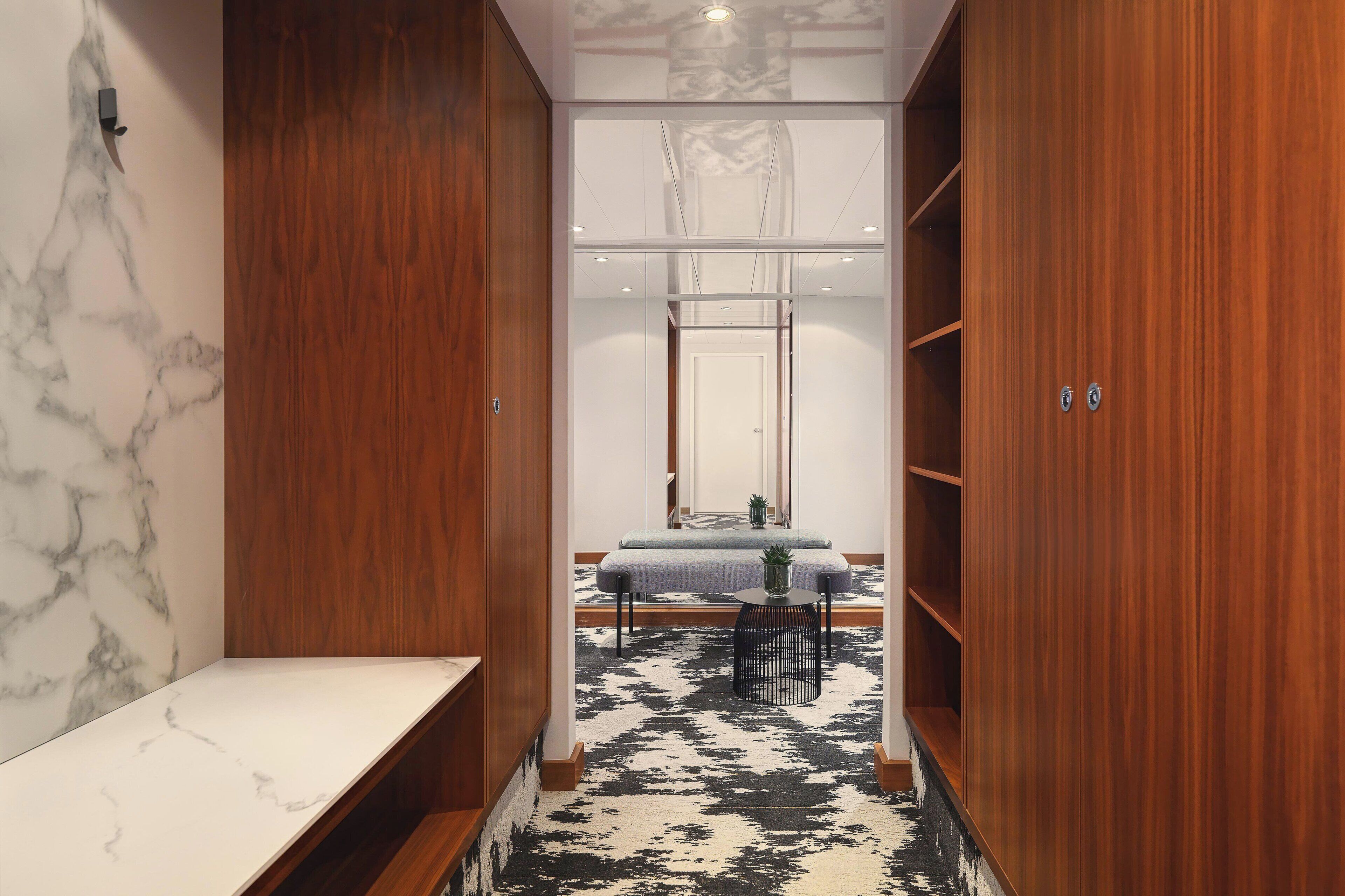A hallway with wooden cabinets, a marble wall, and a black and white patterned rug leading to a seating area with a table and chairs.