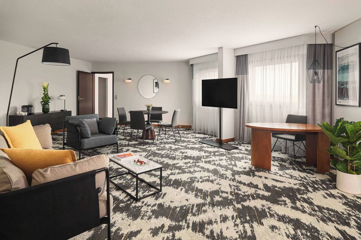 A modern living room with a sofa, coffee table, and dining area. There is a flat-screen TV mounted on the wall and a large window with sheer curtains.