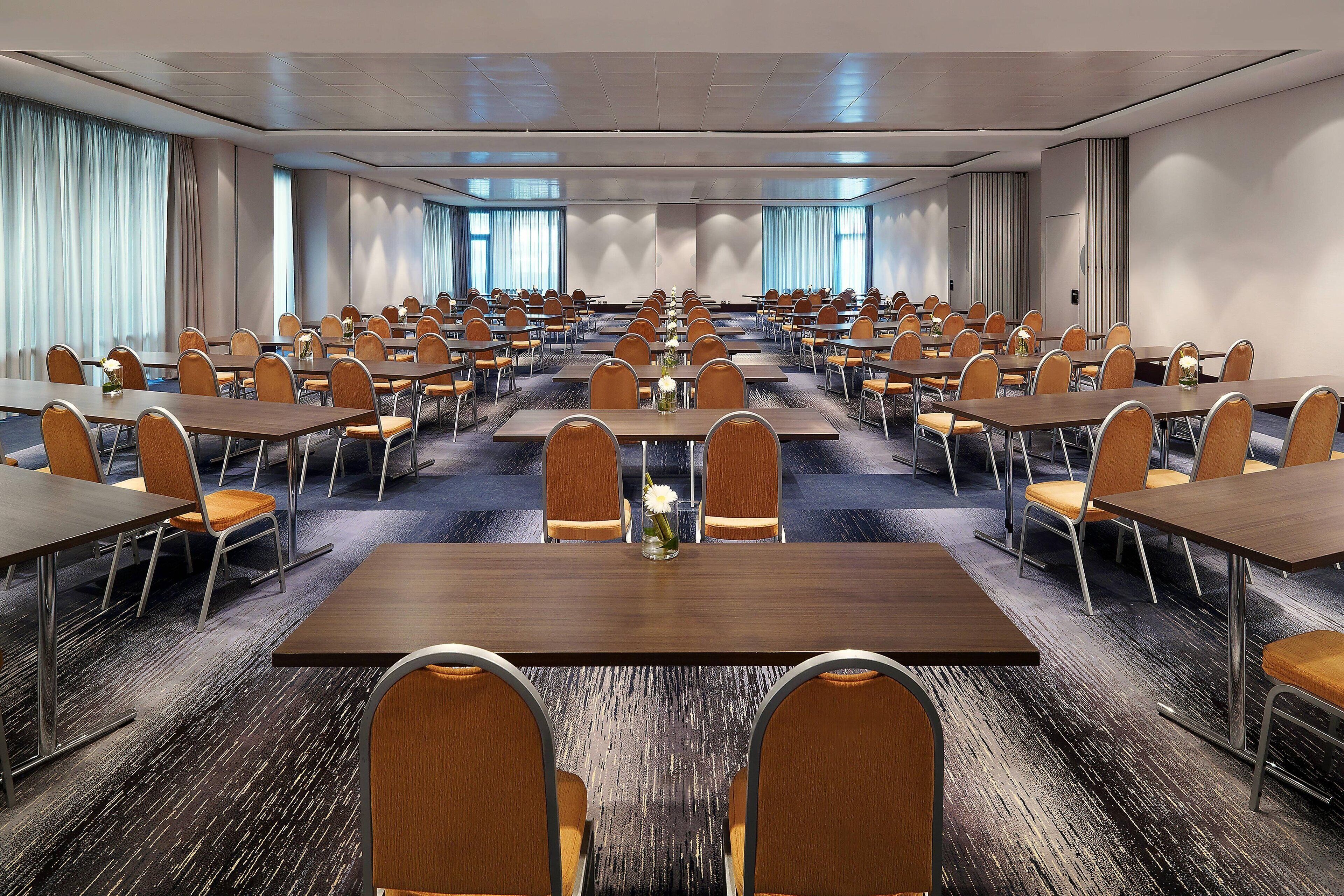 A conference room with multiple tables and chairs, arranged in rows, with a carpeted floor and large windows with curtains.