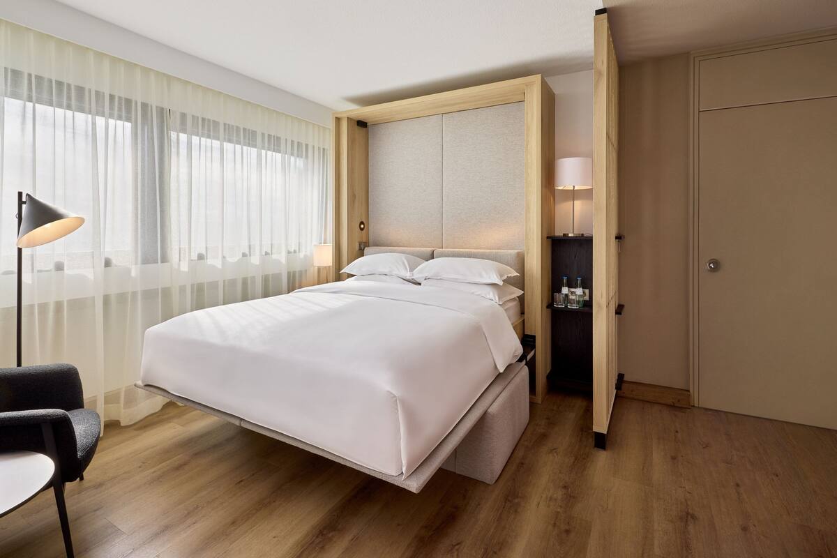 A modern hotel room with a bed, bedside table, lamp, and a view of the outdoors through sheer curtains.