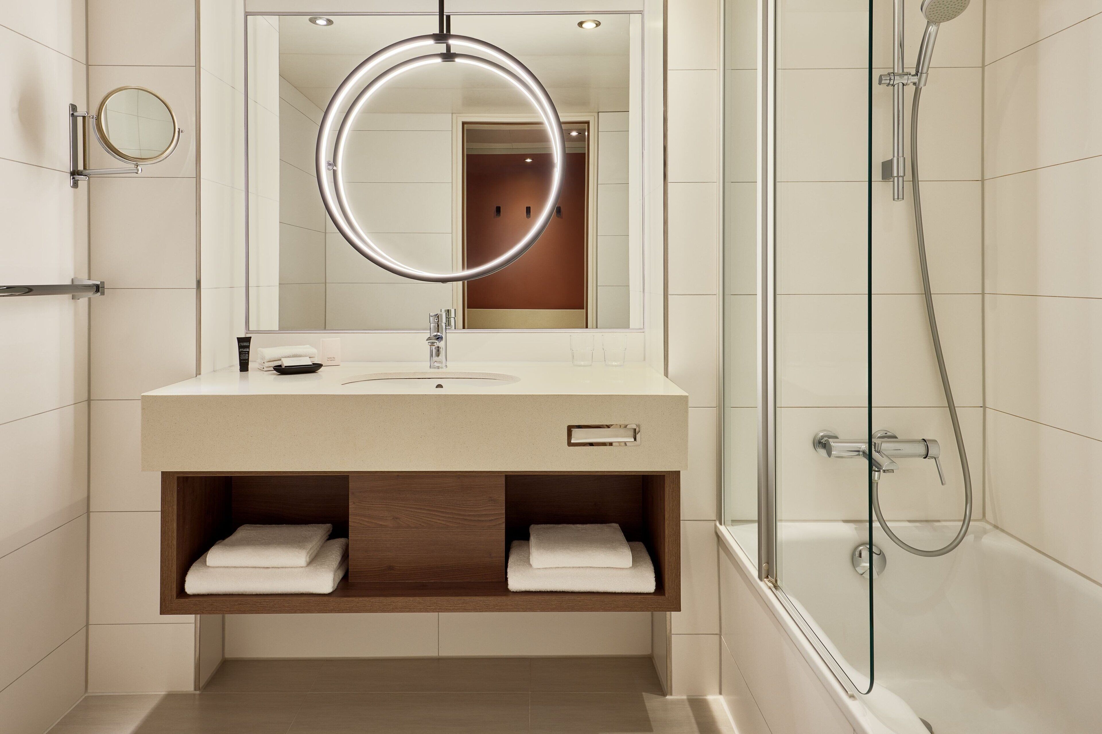 A modern bathroom with a round mirror, a sink, a shower, and wooden shelves with towels.