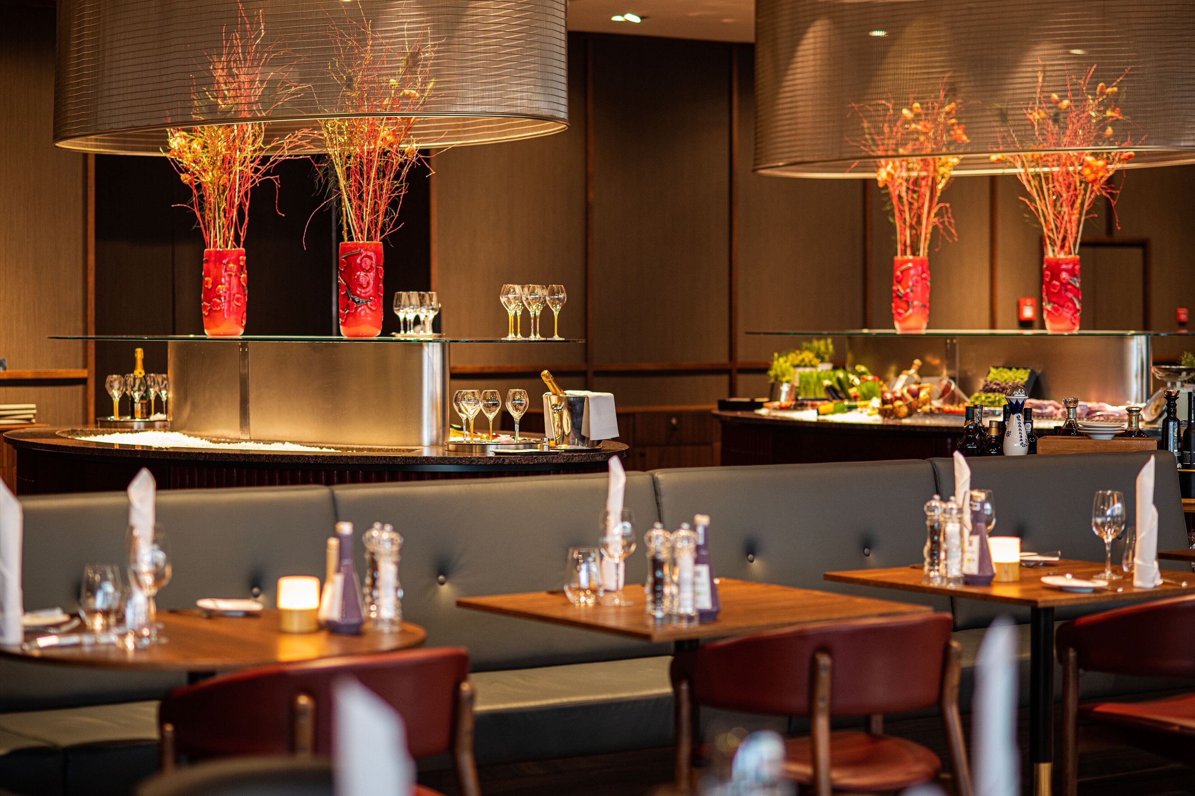 Un restaurant avec des tables dressées pour le service, un coin bar et des vases décoratifs ornés de branches.