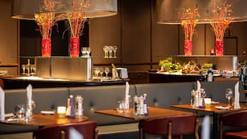 Un restaurant avec des tables dressées pour le service, un coin bar et des vases décoratifs ornés de branches.
