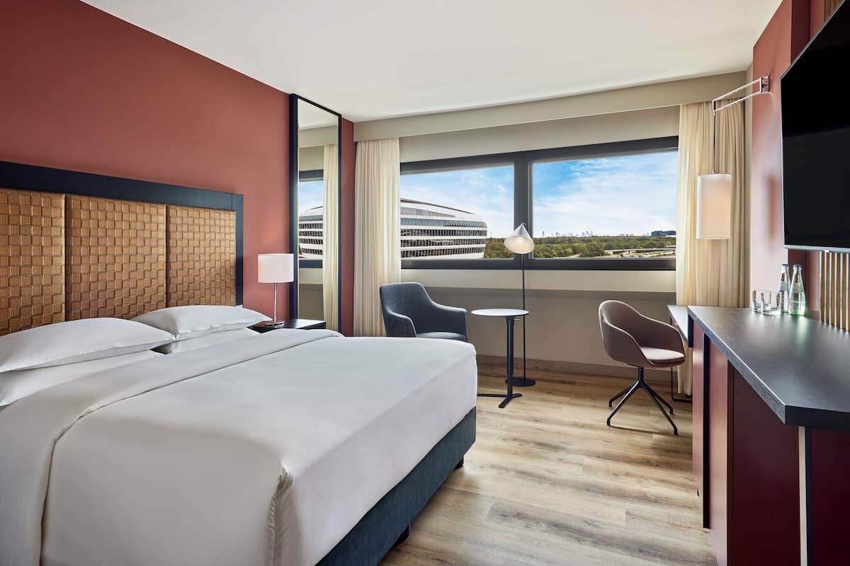 A hotel room with a large bed, a desk, two chairs, a small table, and a view of a building through the window.