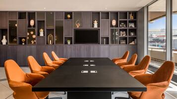 A conference room with a long table, orange chairs, and a dark wood panel wall with shelves containing decorative items.