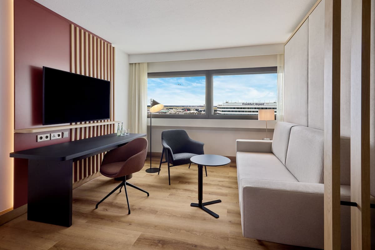 A modern hotel room with a flat-screen TV, a desk, two chairs, a small round table, and a sofa. A large window offers a view of an airport and a building labeled "Frankfurt Airport."