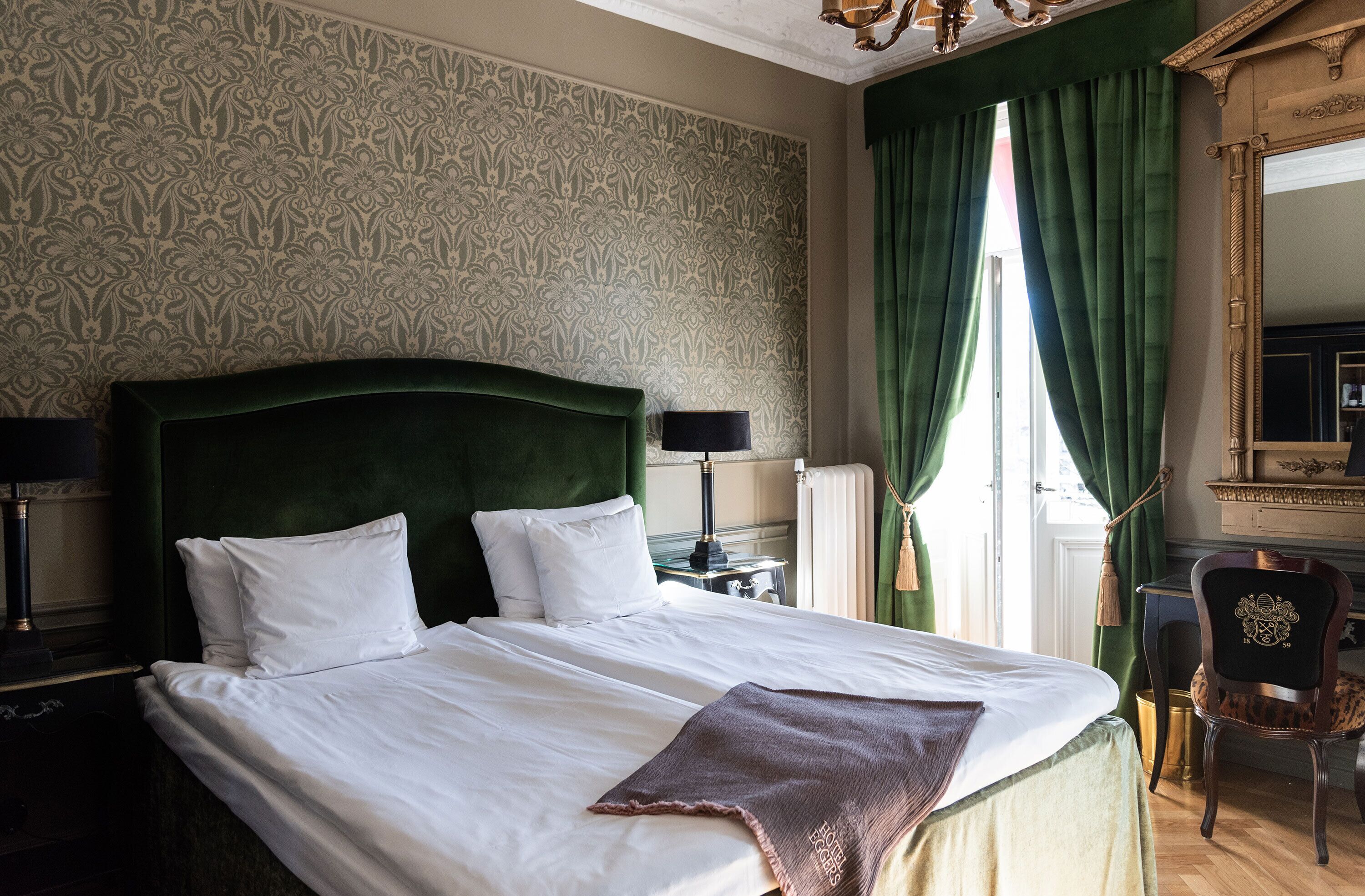 A neatly made bed with white linens and a brown throw, a green upholstered headboard, and a classic lamp beside it.