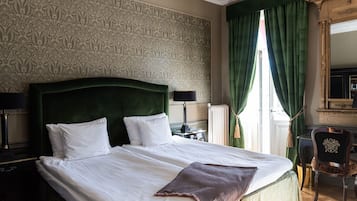 A neatly made bed with white linens and a brown throw, a green upholstered headboard, and a classic lamp beside it.