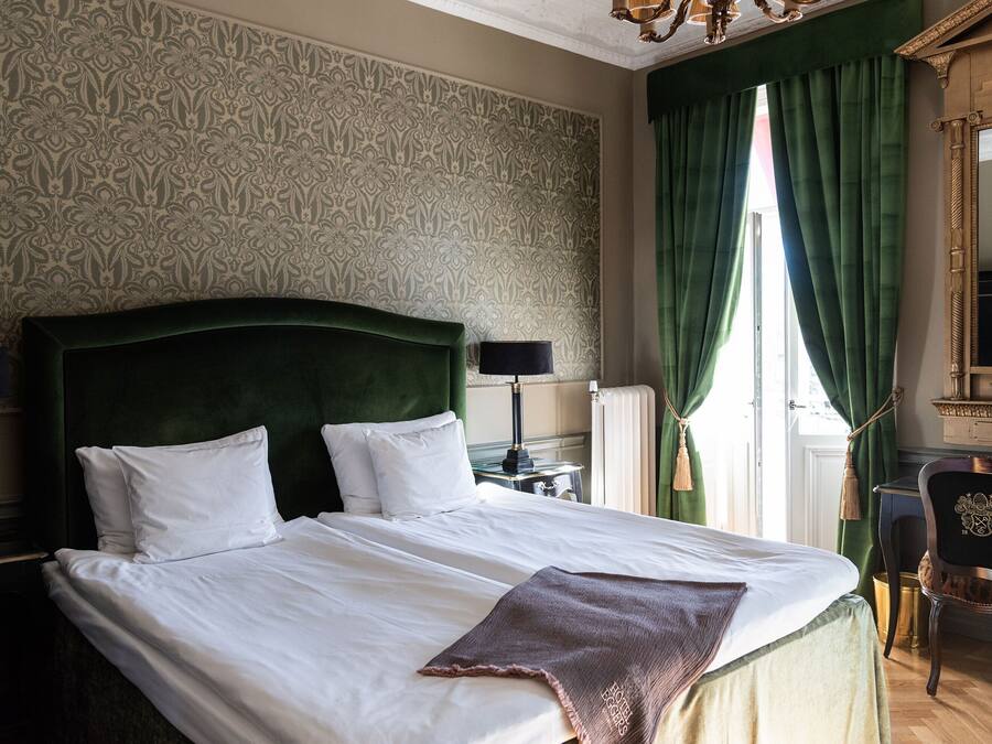 A neatly made bed with white linens and a brown throw, a green upholstered headboard, and a classic lamp beside it.