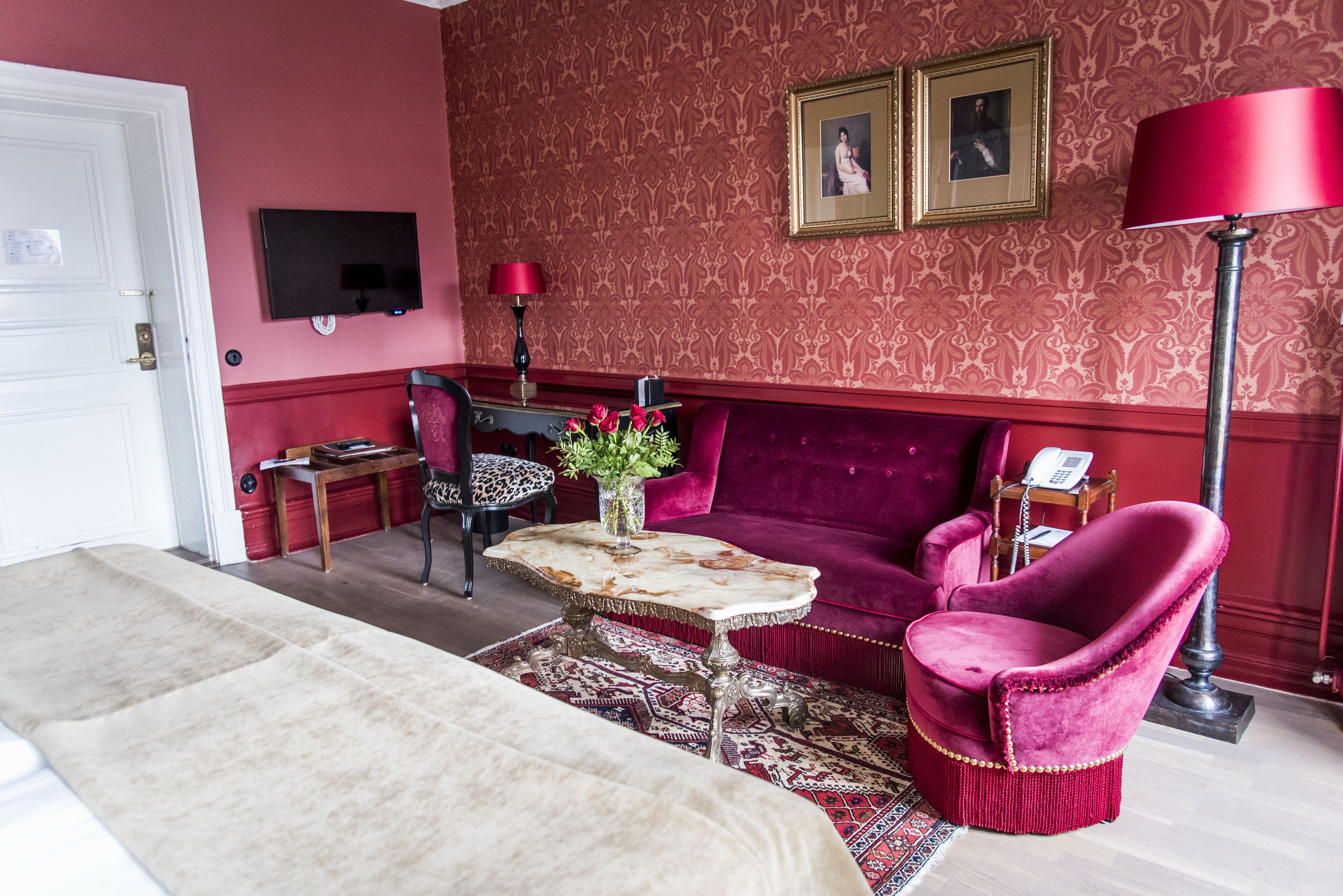 A room with a bed, a red sofa, a table with a vase of flowers, a television on the wall, and a lamp.