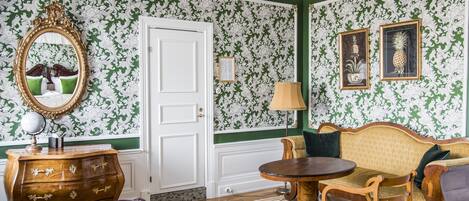 A room with green and white patterned wallpaper, a wooden dresser, a round table, a sofa, a chair, and a mirror with a gold frame.