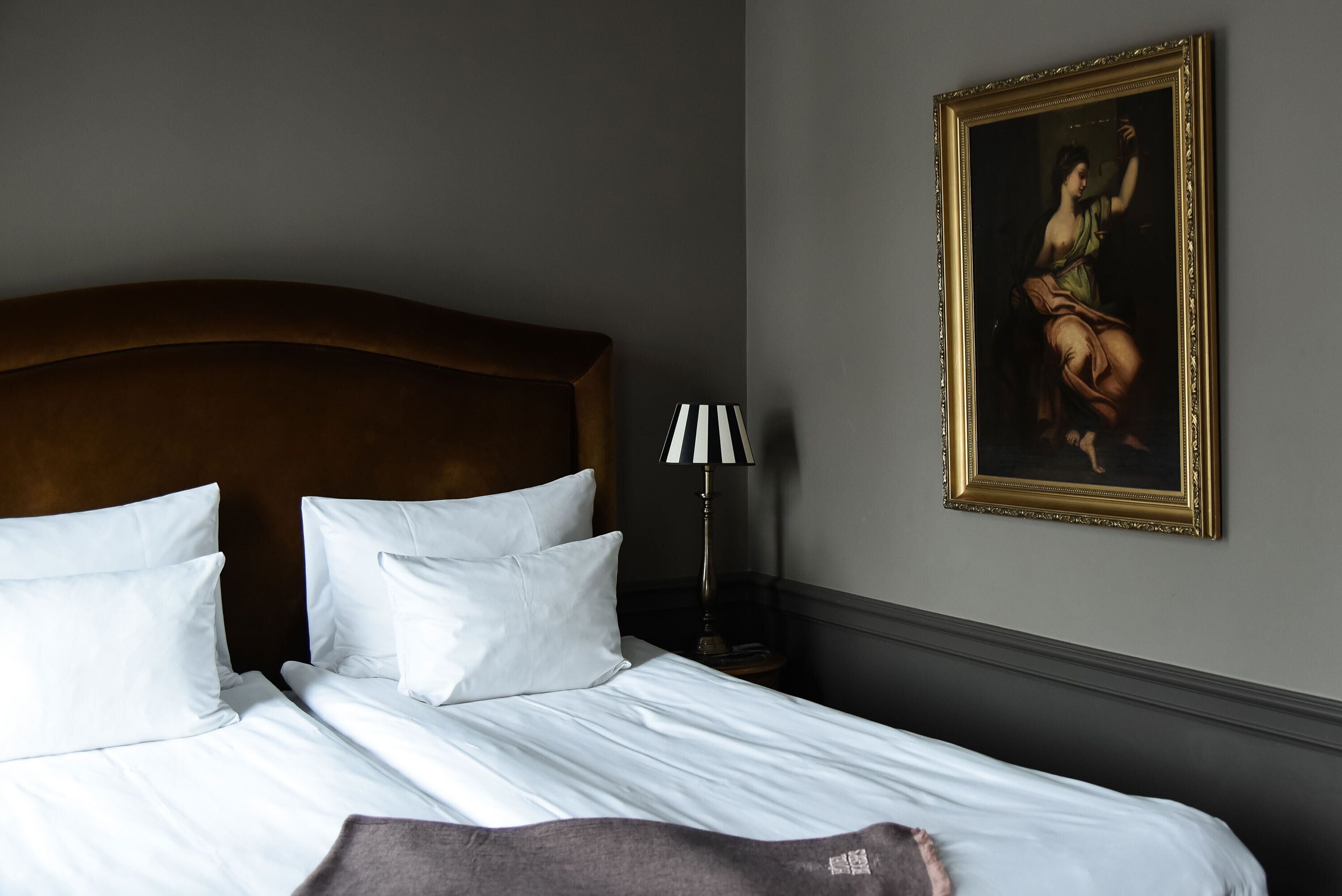 A neatly made bed with white linens and pillows, a wooden headboard, a lamp with a striped shade, and a framed painting on the wall.
