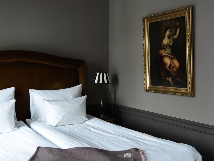 Una cama bien hecha con sábanas y almohadas blancas, un cabecero de madera, una lámpara con pantalla a rayas y un cuadro en la pared.