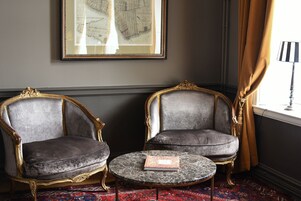 Dos sillones ornamentados con una mesa de mármol en el centro, un mapa enmarcado en la pared y una ventana con cortinas.