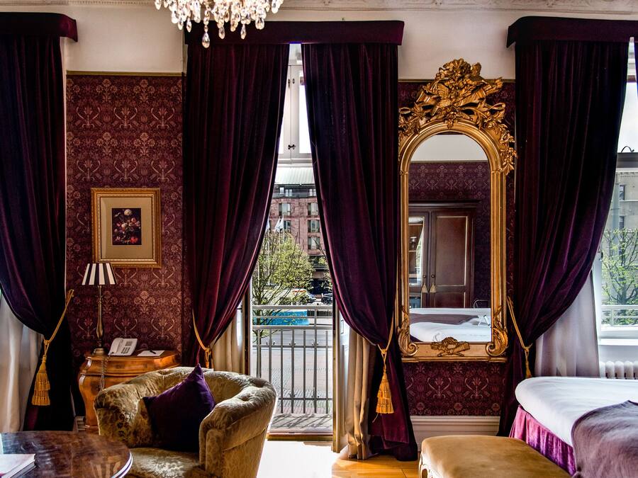 A room with a large ornate mirror, a plush armchair, a small table with a lamp, and a view of the street through a window with dark curtains.