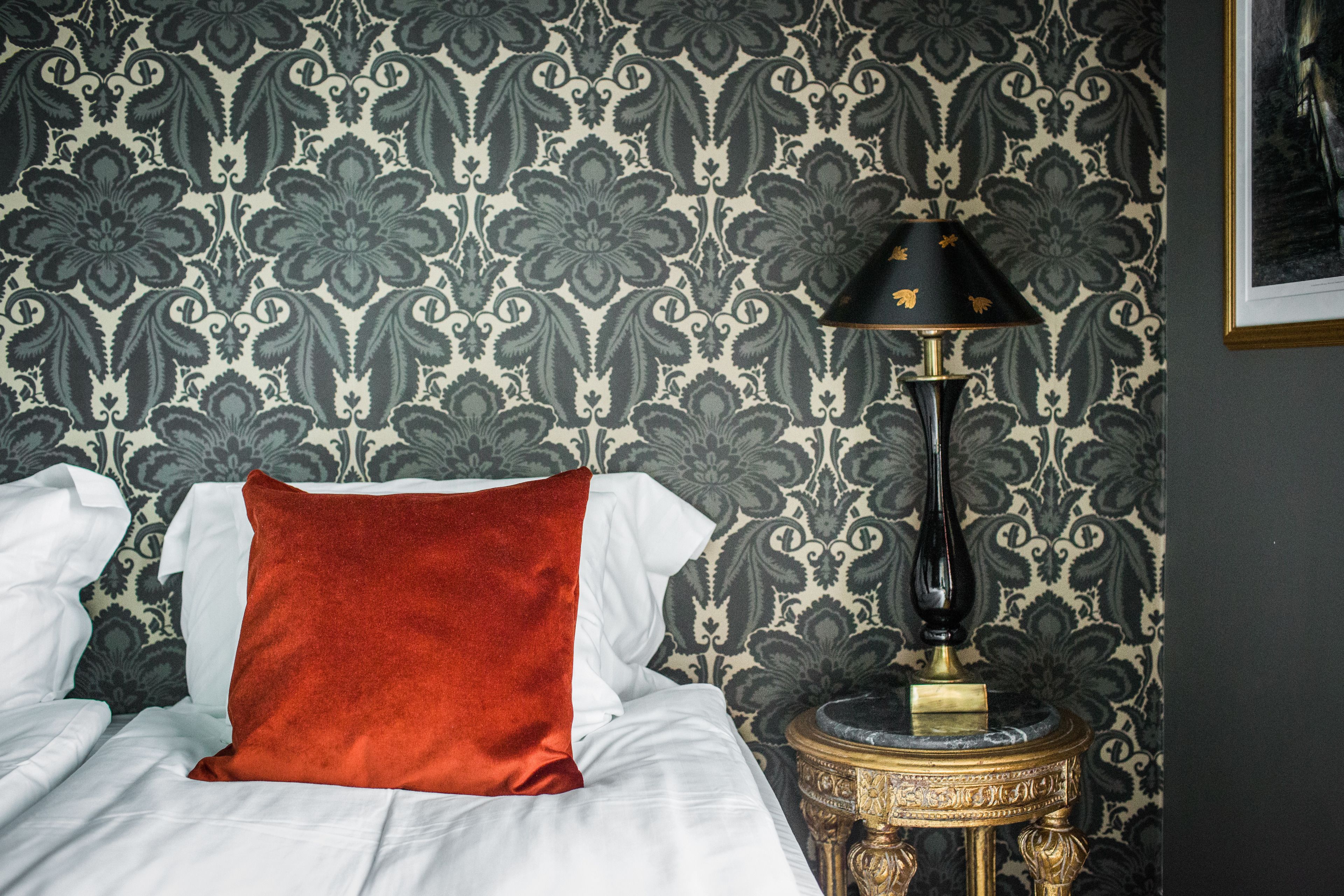 A neatly made bed with a red pillow, a gold-framed lamp, and a patterned wallpaper.