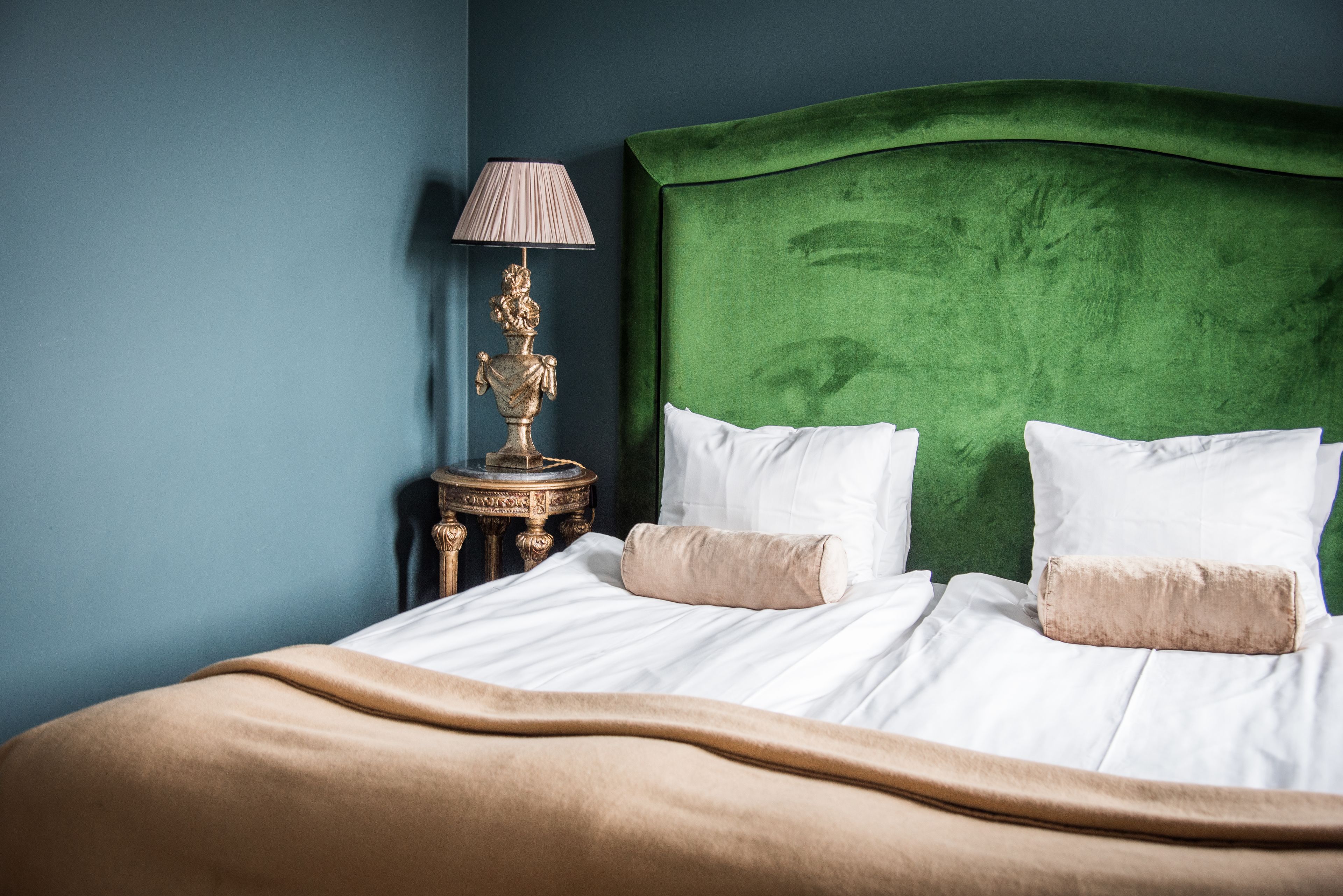 A neatly made bed with white linens and two beige pillows, a green upholstered headboard, and a gold lamp on a matching side table.