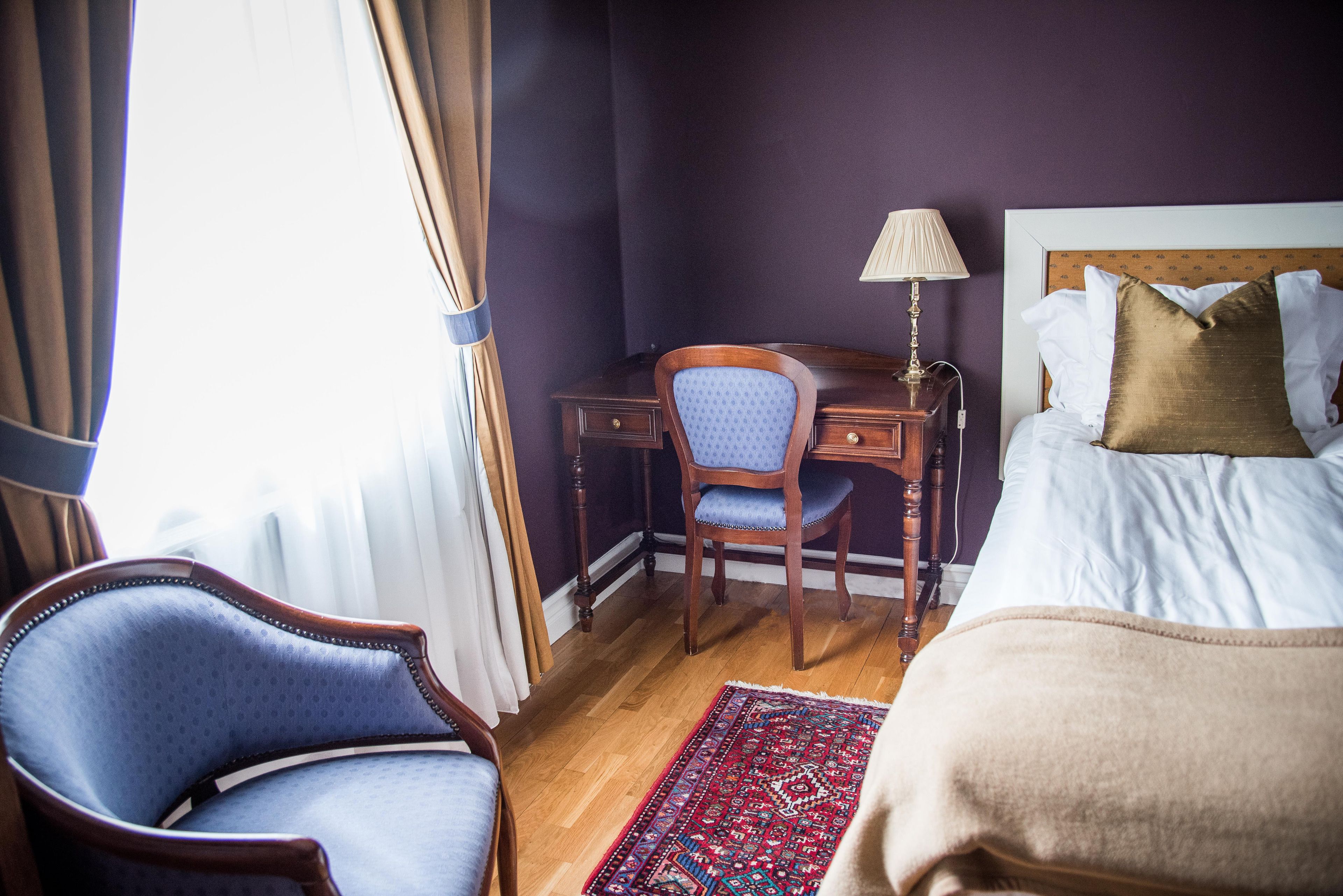 A hotel room with a bed, a desk with a chair, a lamp, and a patterned rug.