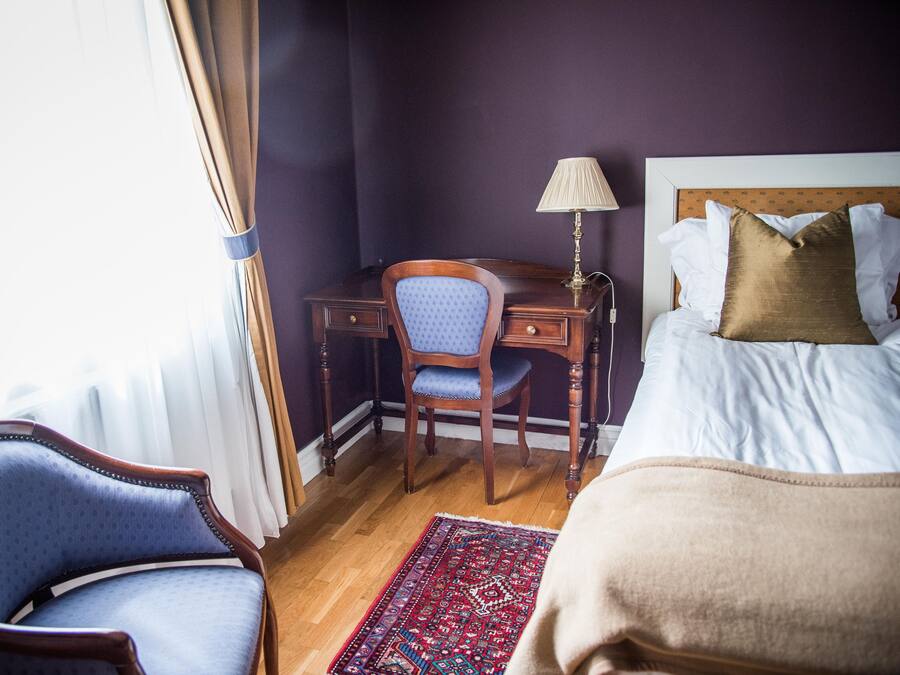 A hotel room with a bed, a desk with a chair, a lamp, and a patterned rug.