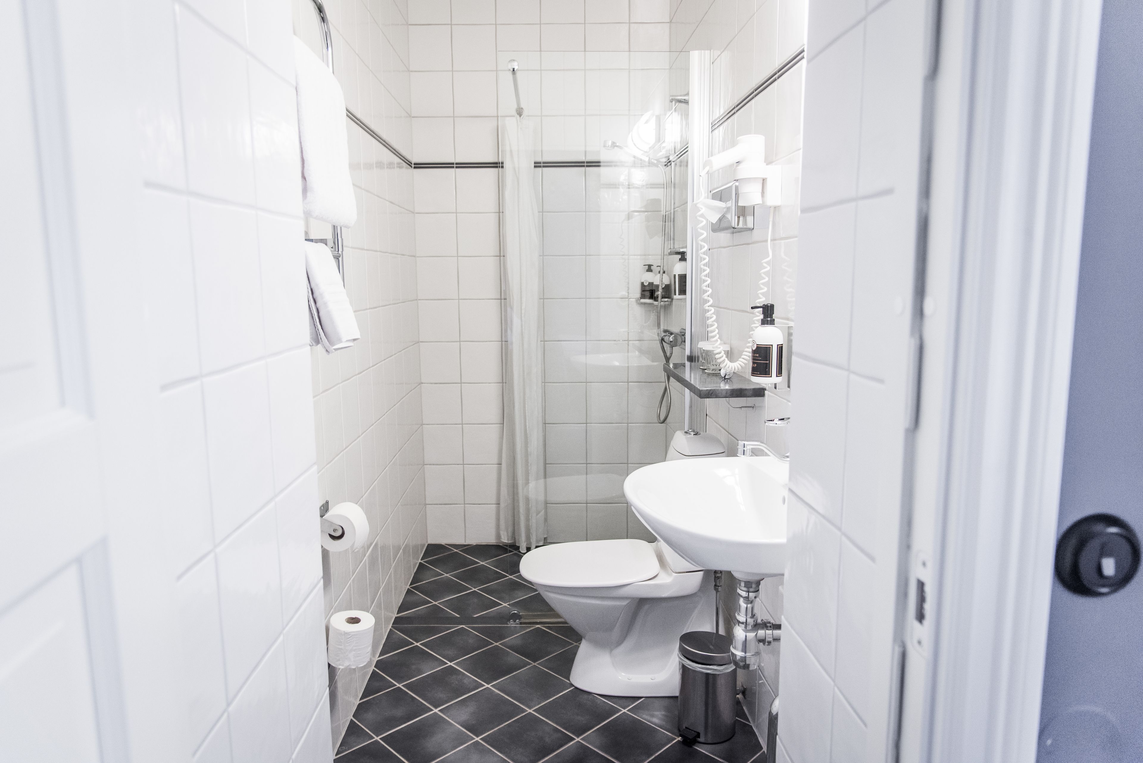 Un baño pequeño y moderno con azulejos blancos, suelo de baldosas negras, inodoro blanco, lavamanos y ducha.