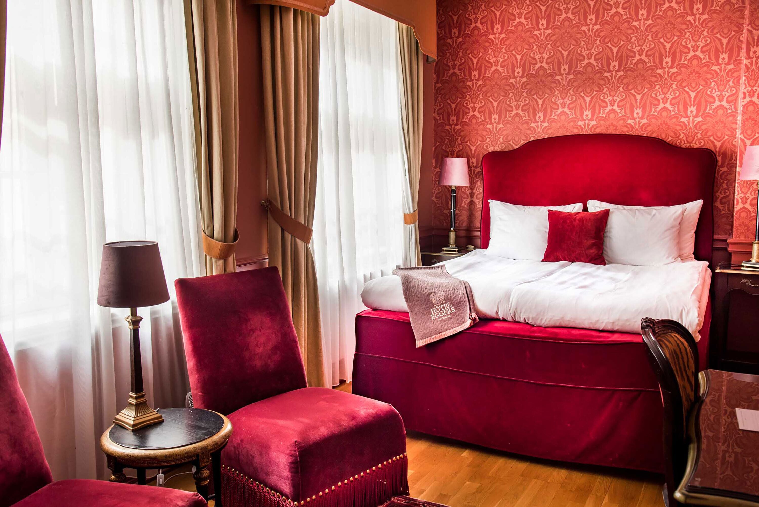 Een hotelkamer met een rode bekleden bed, een rode fauteuil, een klein rond tafeltje met een lamp en behang met een patroon.