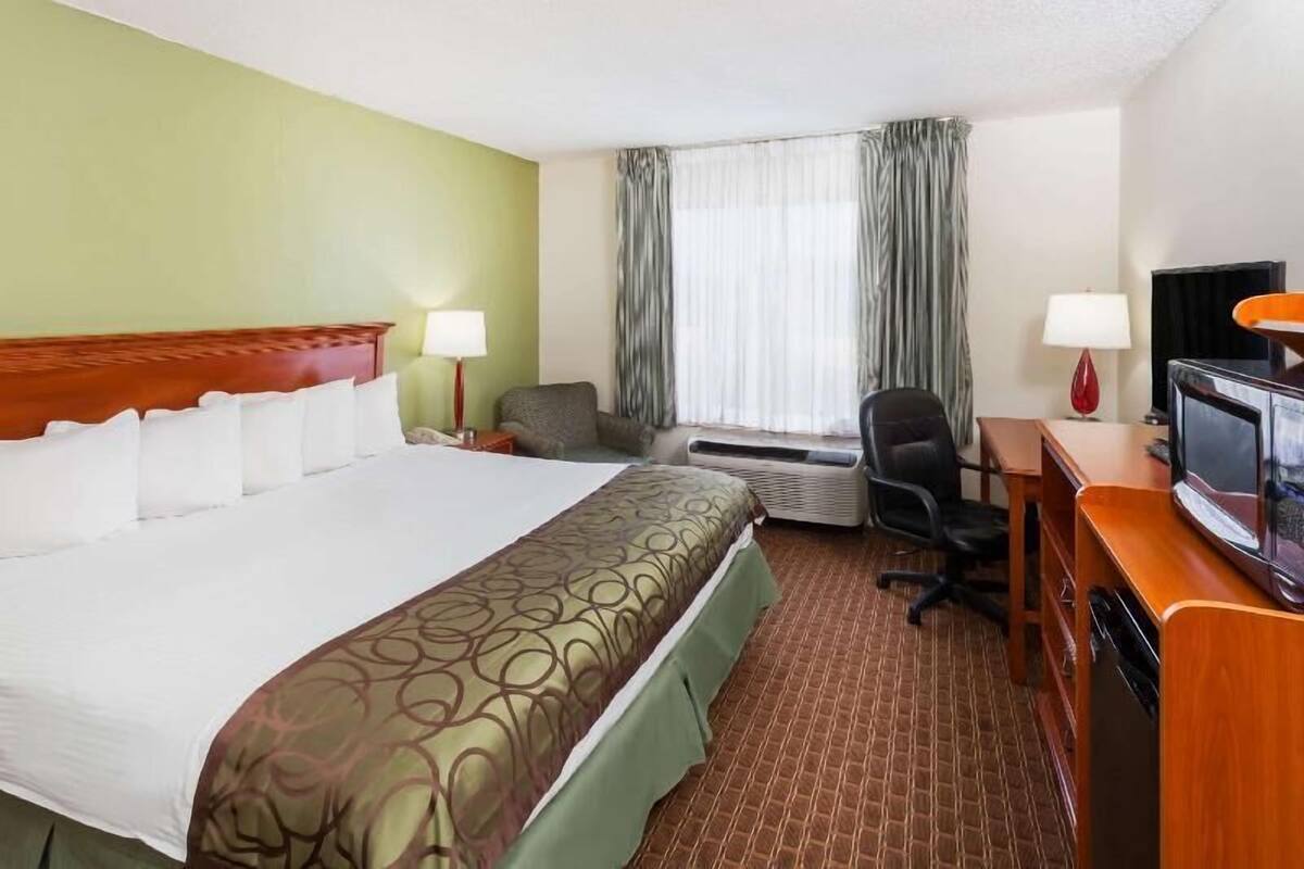 A hotel room with a bed, a desk with a television, an air conditioning unit, and a chair.