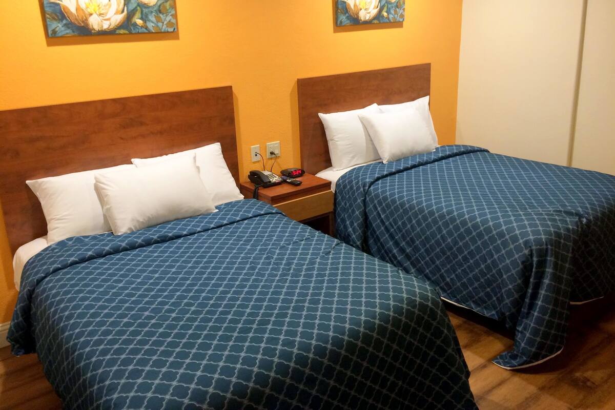 A hotel room with two beds, each with a blue patterned bedspread and white pillows. There are two paintings of flowers on the walls, and a nightstand with a phone and a clock displaying 5:24.