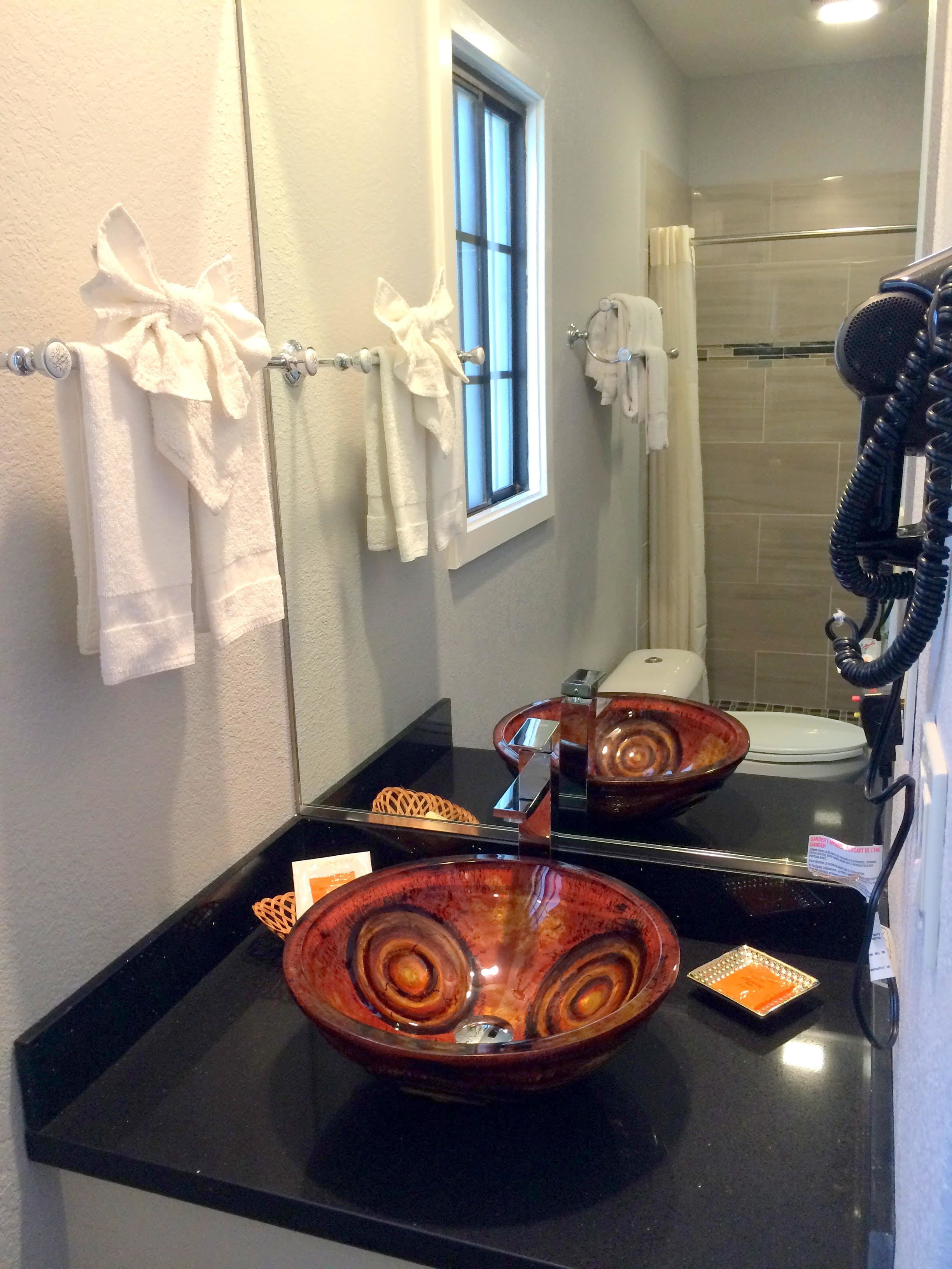 Un baño con encimera negra, un lavabo de vidrio, un perchero con toallas blancas, una ventana y una zona de ducha.